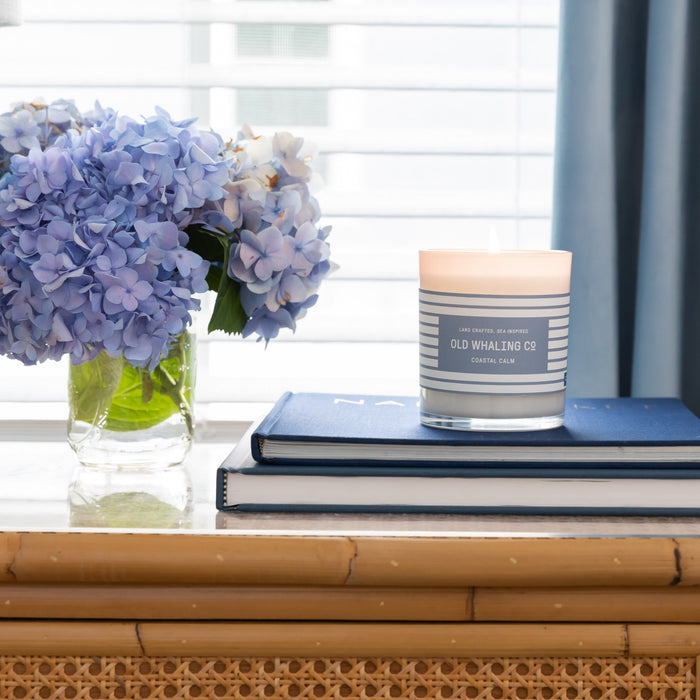 Coastal Calm Candle with lit, glowing wick, sitting on stack of books beside hydrangea arrangement